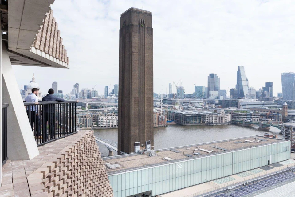 A tall brown tower with a cityscape view in the background, seen from a building's balcony overlooking a river.