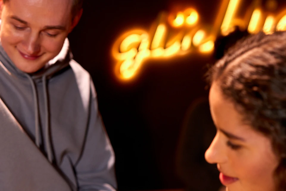 Two people seated indoors under warm lighting, one holding a utensil while the other smiles. A neon light is in the background.