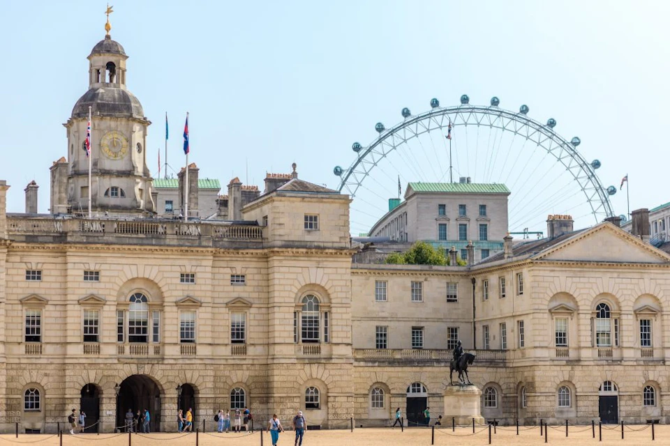 Historic building with an equestrian statue in front, visitors nearby. Large Ferris wheel and modern buildings in the background.