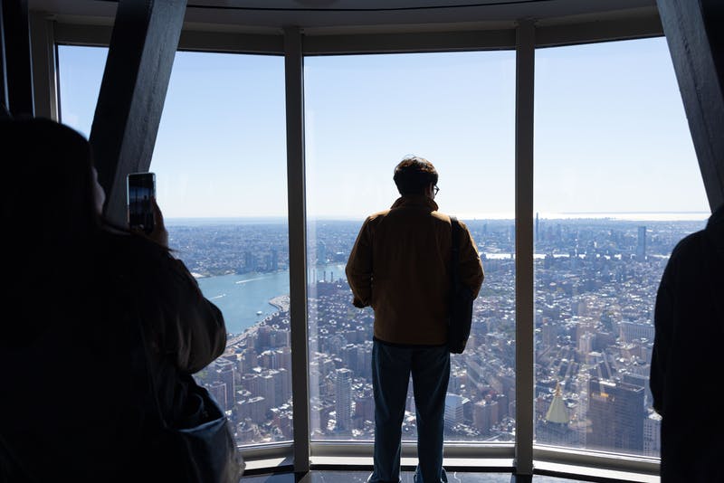 Ένα άτομο στέκεται σε ένα ψηλό παράθυρο με θέα στον ορίζοντα της πόλης και ένα άλλο άτομο φωτογραφίζει τη σκηνή.