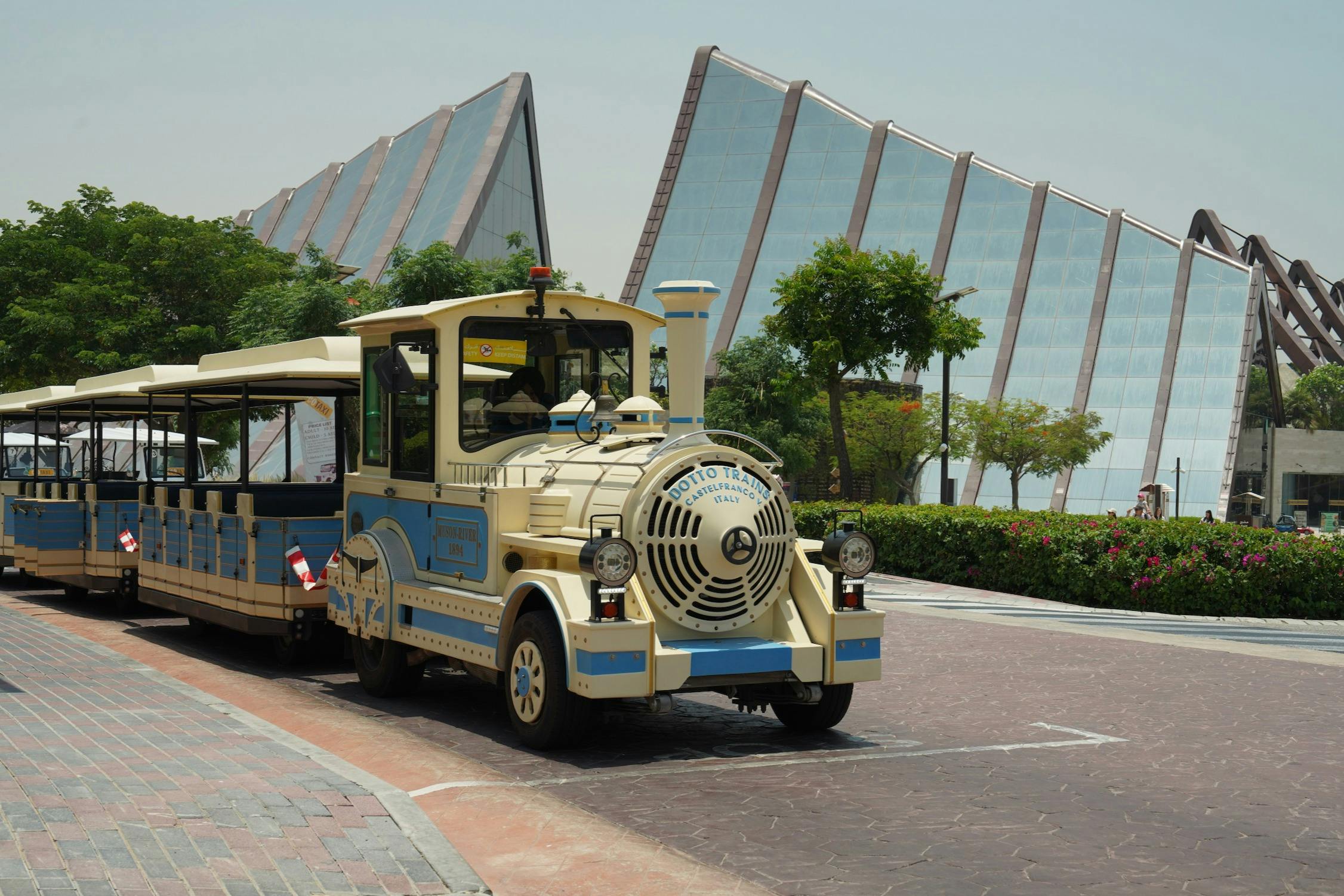 A blue and white tour train parked in front of modern glass buildings and greenery on a sunny day.