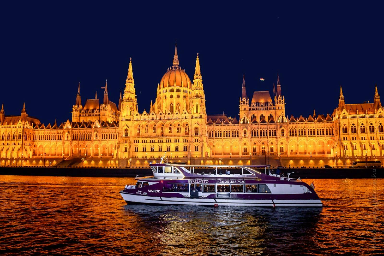 Eine beleuchtete Bootstour auf einem Fluss vor einem beleuchteten, verschnörkelten Gebäude bei Nacht.
