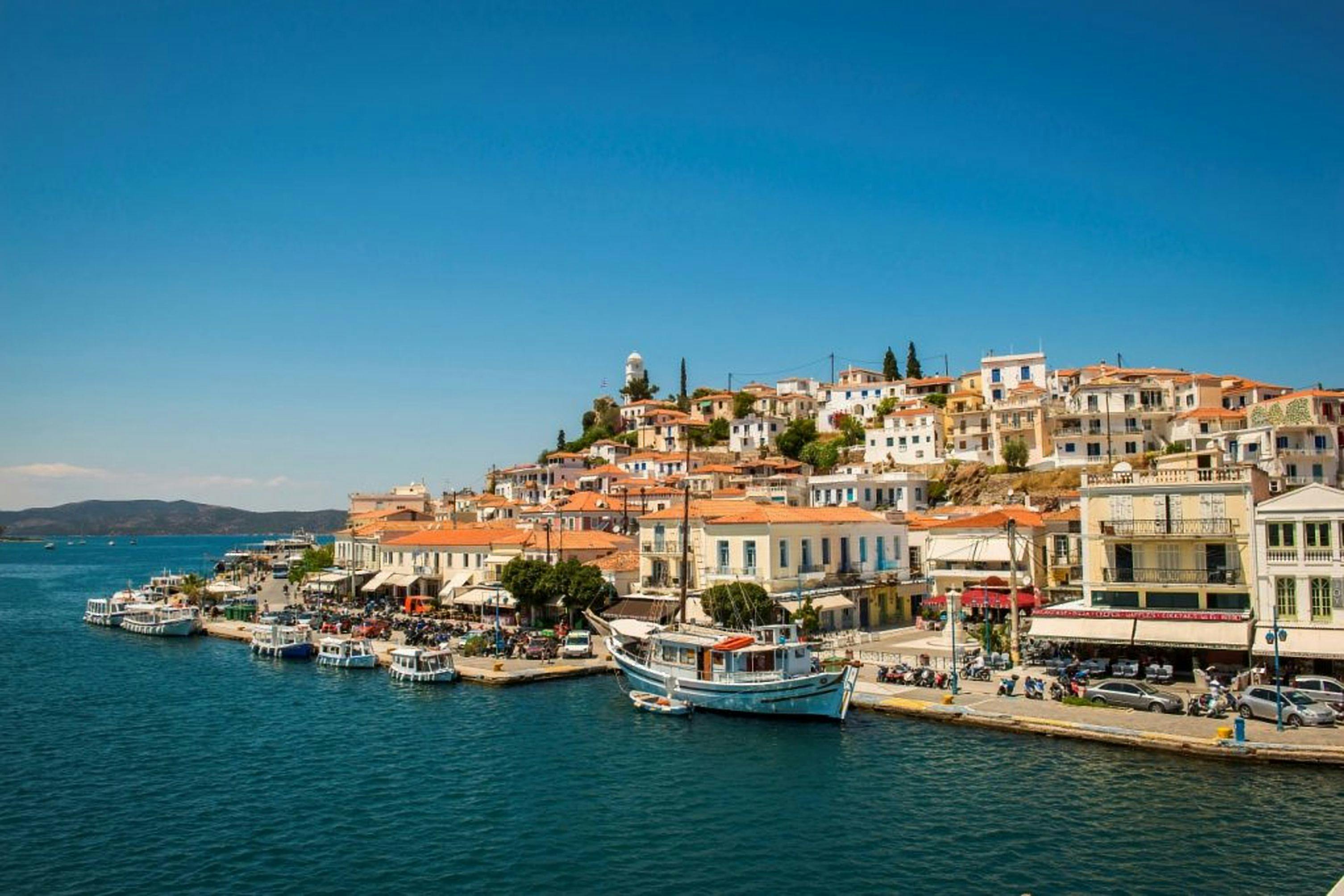 Прибрежный город с белыми и пастельными зданиями, лодками, пришвартованными на набережной, и склоном холма с большим количеством домов под чистым голубым небом.