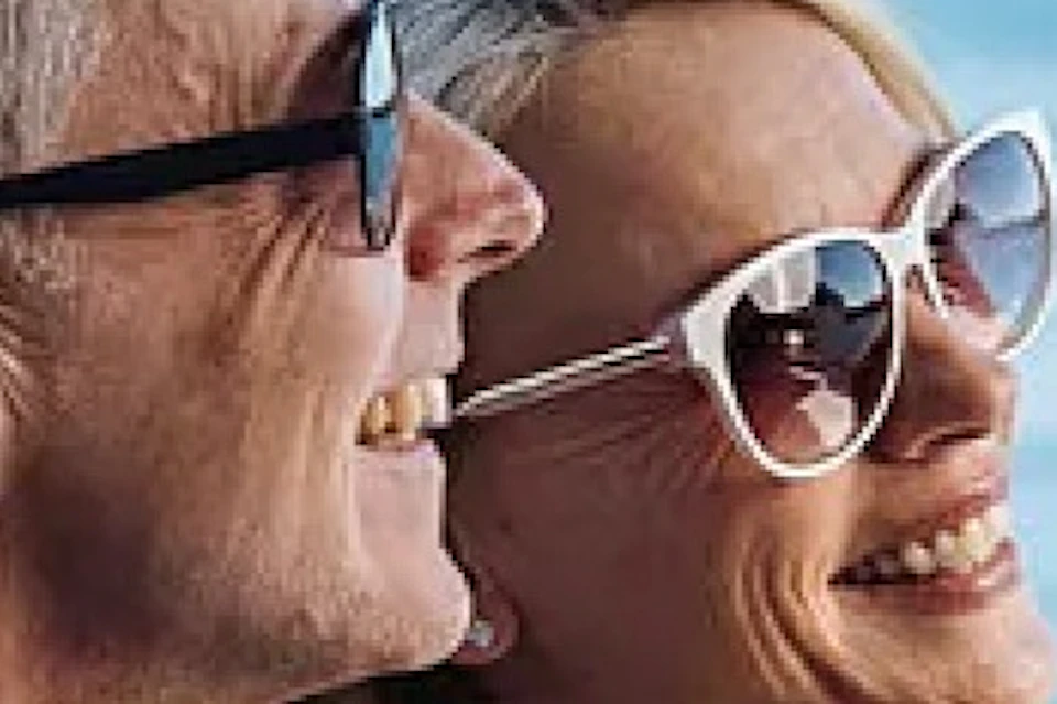 Two people smiling on a boat with a city skyline and water visible in the background under a clear sky.