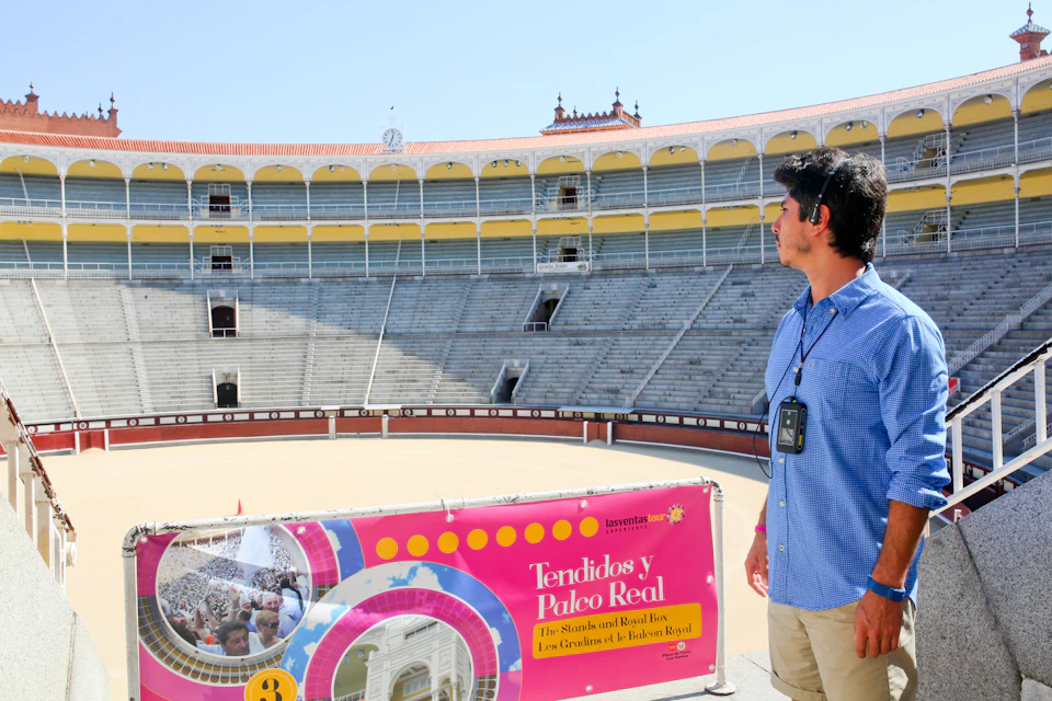 A man with an audio guide stands in an empty bullring, looking at the seats. A pink sign reads "Tendidos y Palco Real."