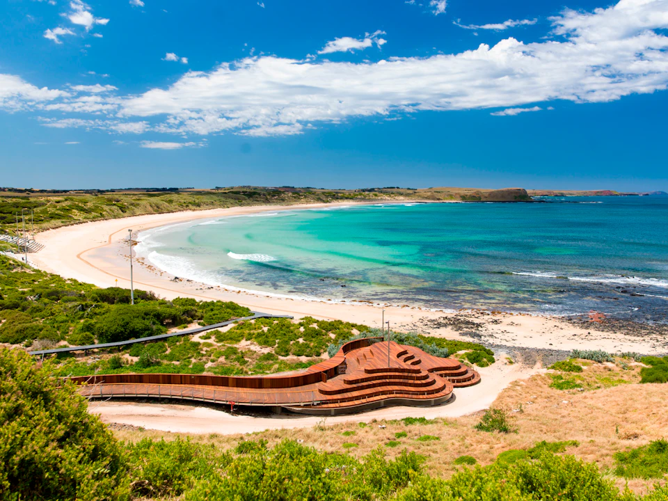 A scenic coastal view with a wooden observation deck, green bushes, a sandy beach, and turquoise ocean waves under a cloudy sky.