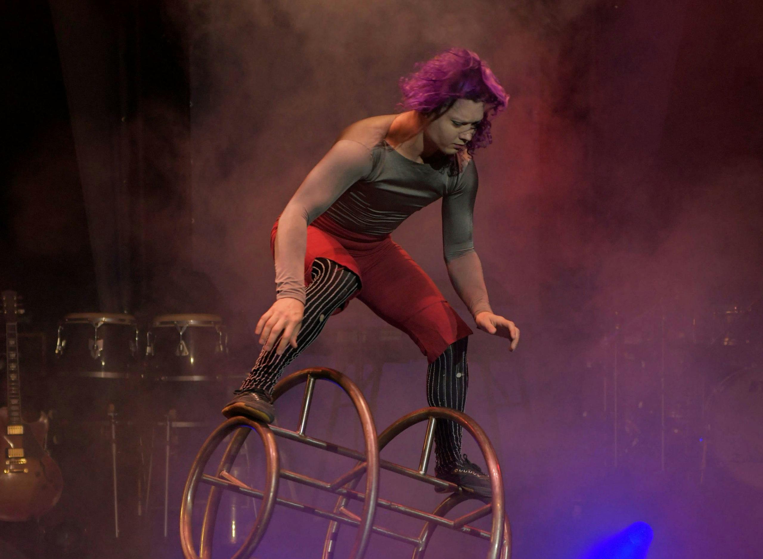 Artist med lila hår balanserar på en cirkushjulsapparat på en rökig scen, trumset och gitarr i bakgrunden.