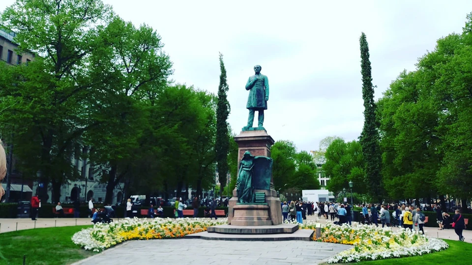 Bronze statue of a man on a pedestal in a park, surrounded by blooming flowers and trees, with people walking nearby.
