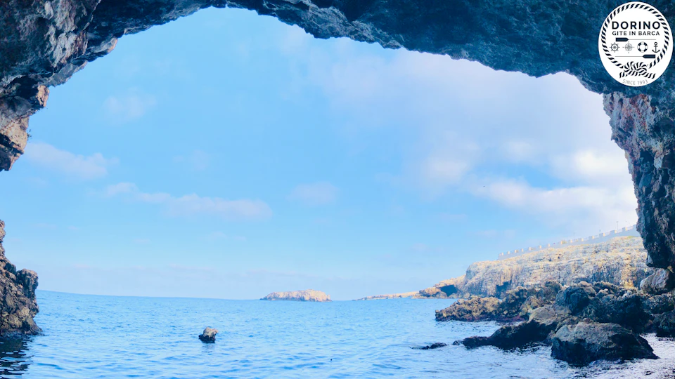 View from inside a cave looking out to a calm sea, with rocky cliffs and small islands in the distance under a clear blue sky.