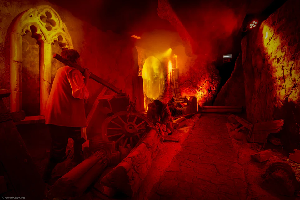 A dimly lit, red-hued room with people interacting with machinery and stone structures, evoking a dramatic atmosphere.