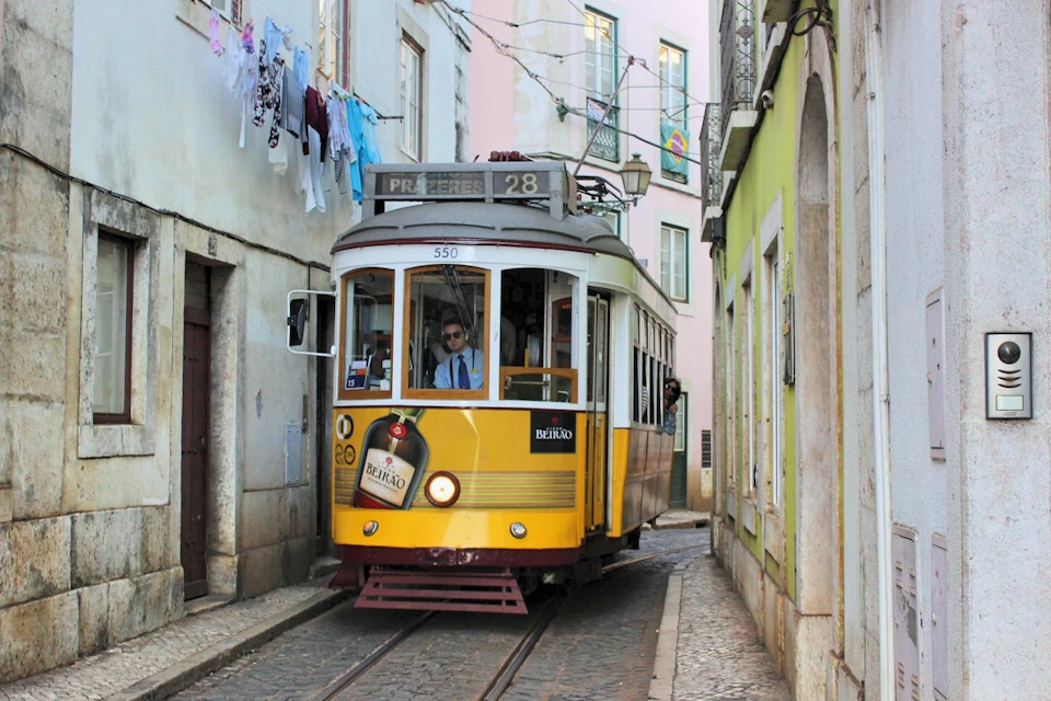 Yellow tram traveling through a narrow street lined with pastel buildings, with clothes hanging on a line above.