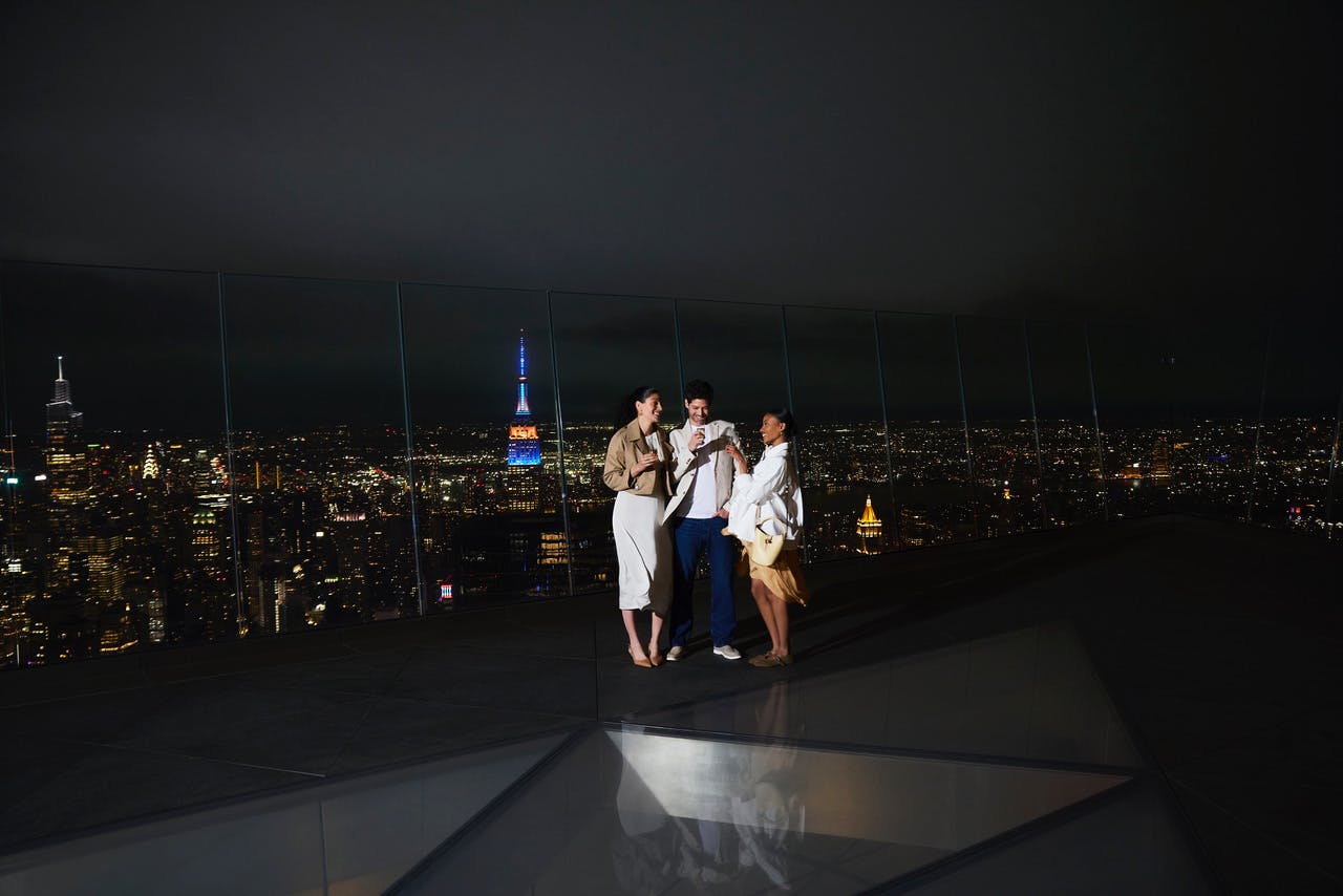 Tři lidé stojí v noci na střešní vyhlídkové plošině a za nimi panorama města s Empire State Building.