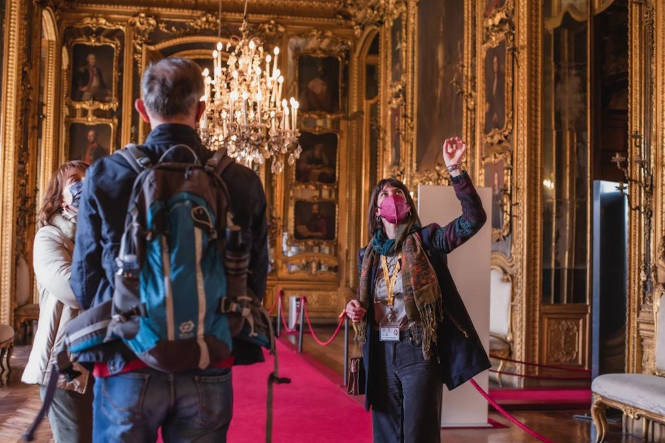 A tour guide gestures inside an ornate room with chandeliers and portraits, while a visitor with a backpack listens attentively.