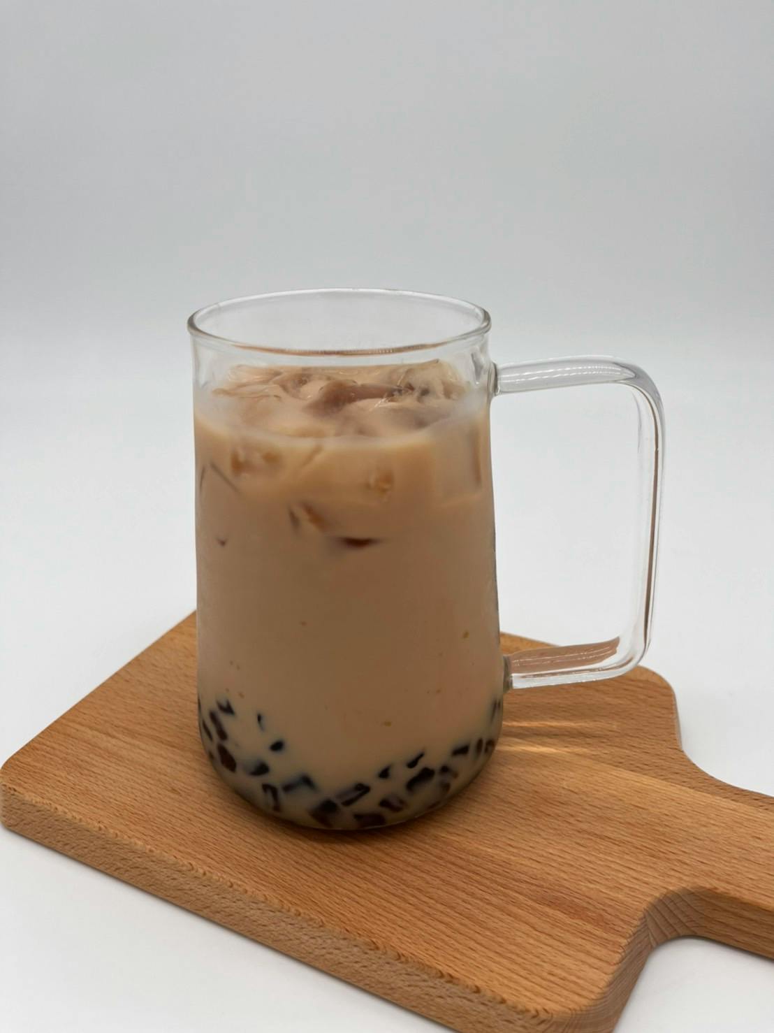 A glass mug of iced milk tea with tapioca pearls, placed on a wooden board against a light background.
