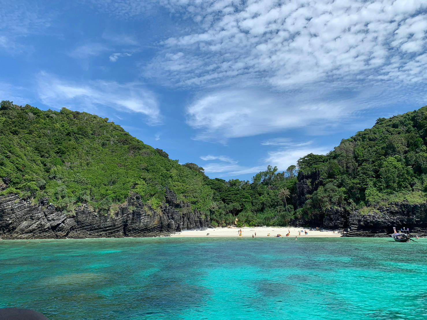 Une plage immaculée avec des collines verdoyantes, une eau claire et turquoise, et quelques personnes qui se détendent sous un ciel partiellement nuageux.