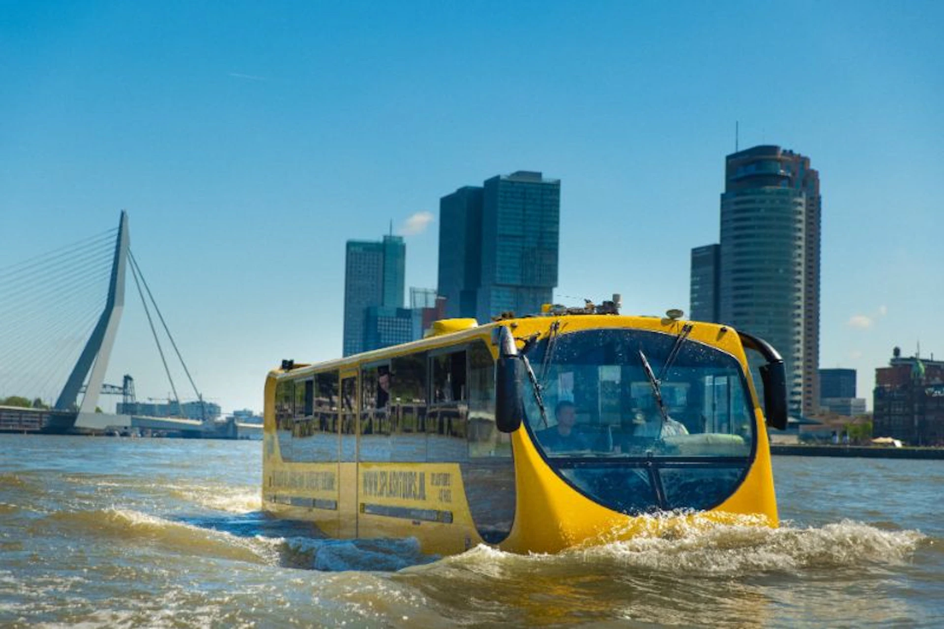 A yellow amphibious bus travels on water in front of a cityscape with modern skyscrapers under a clear blue sky.