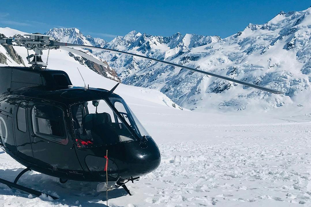 A black helicopter parked on a snow-covered mountain with a backdrop of snowy peaks under a bright blue sky.