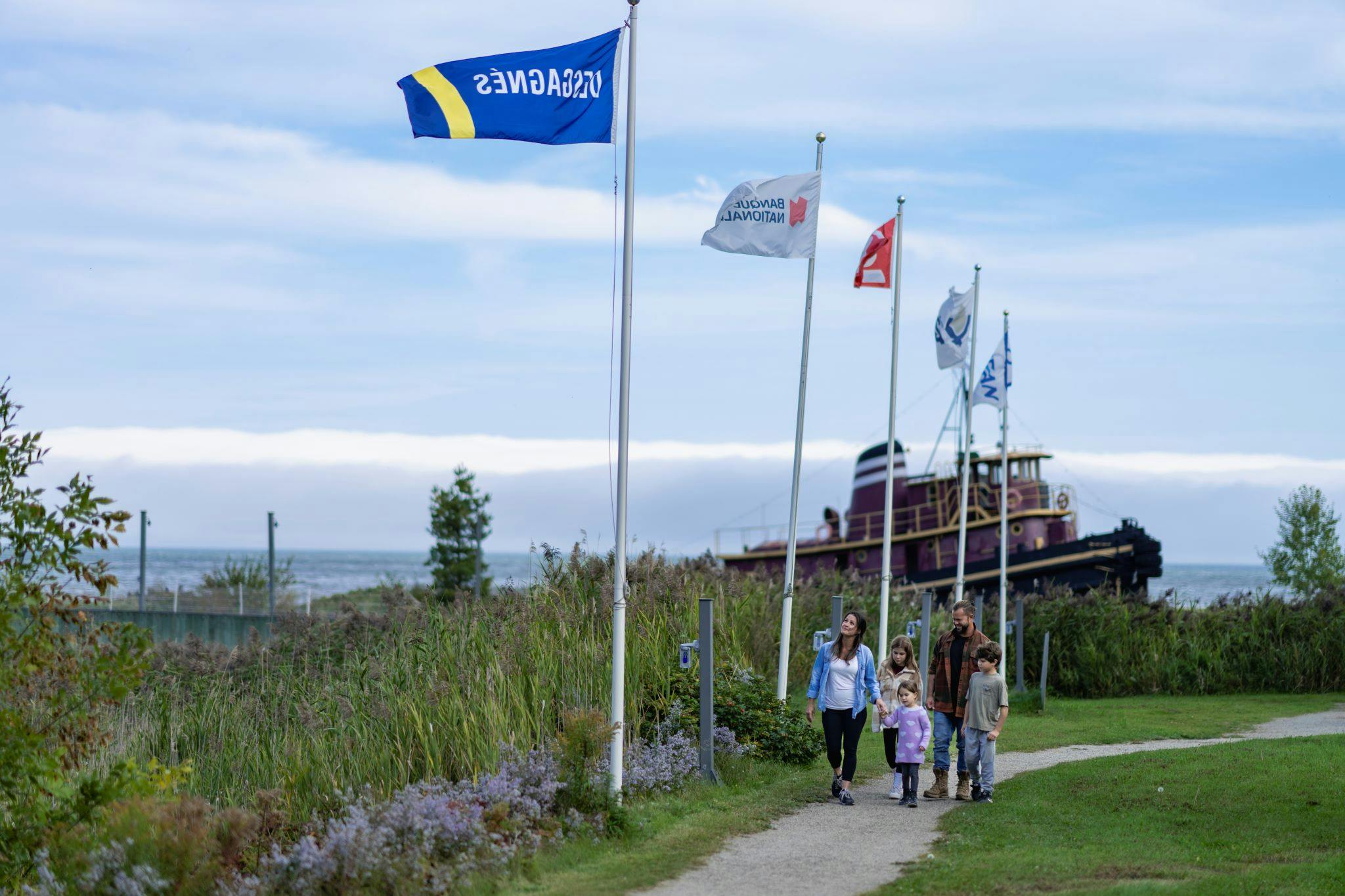En familie går på en sti ved siden af højt græs og flag med en gammel båd i baggrunden under en delvist overskyet himmel.