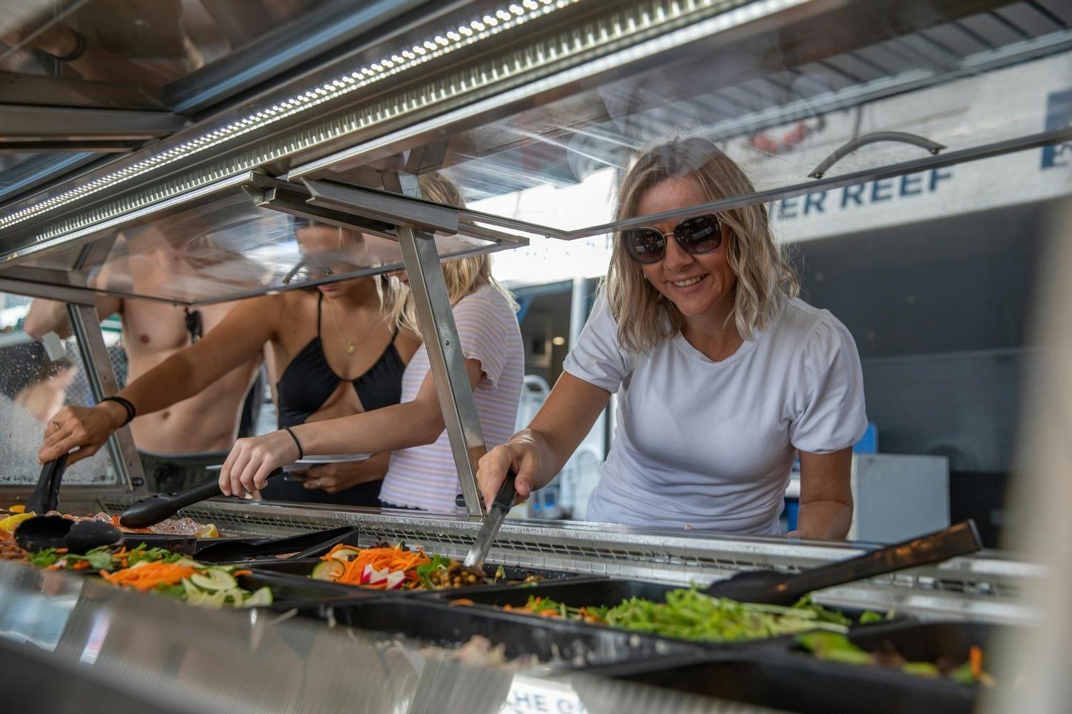 Eine lächelnde Frau mit Sonnenbrille bedient sich an einer Salatbar, in deren Schalen verschiedene frische Gemüsesorten zu sehen sind.