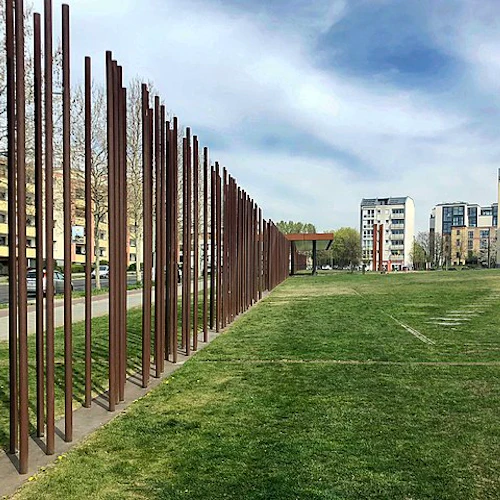 Höga rostfärgade metallstänger kantar en trottoar bredvid ett gräsbevuxet område med flerbostadshus i bakgrunden.