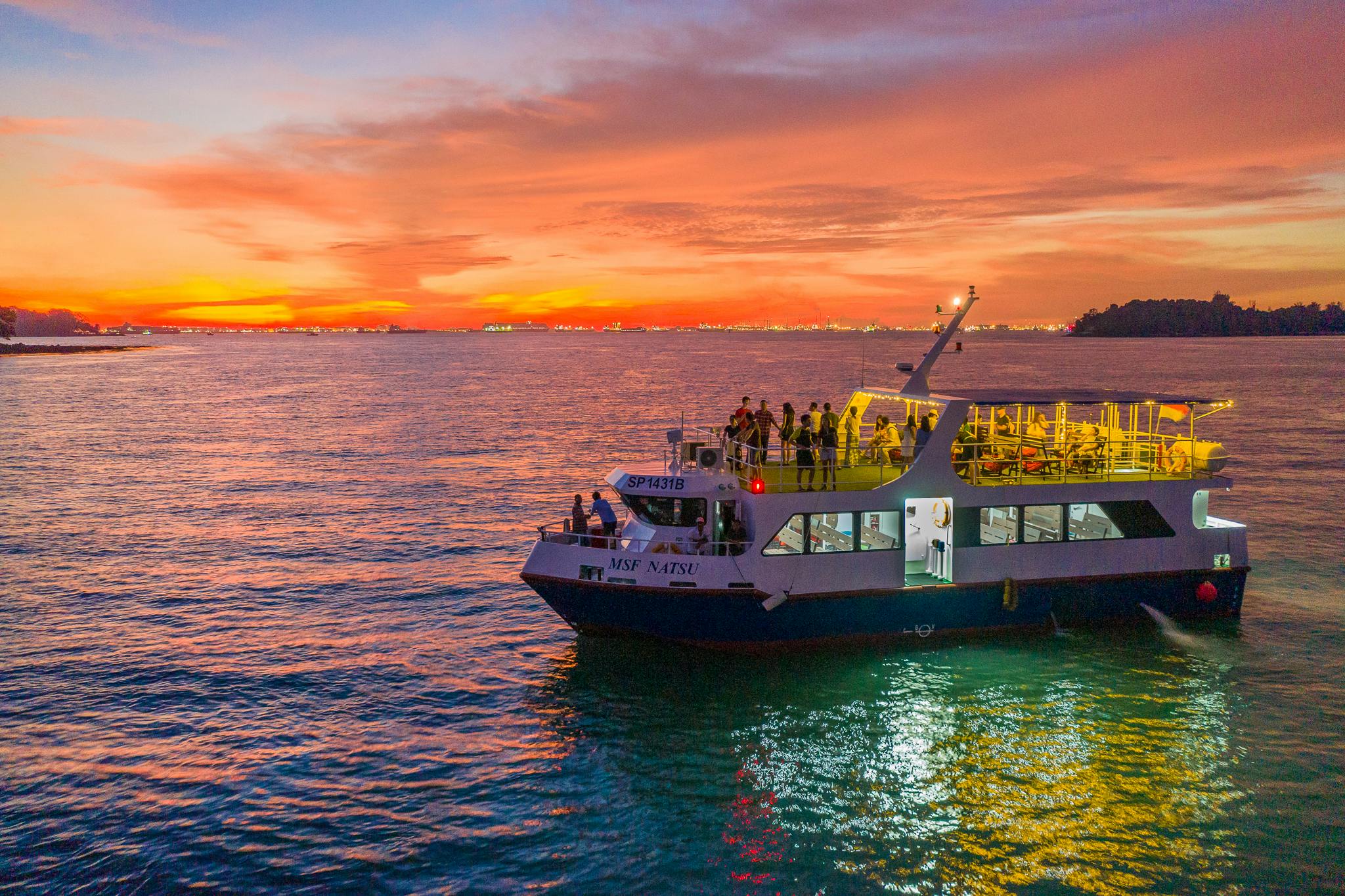 A ferry with passengers on a calm body of water at sunset, with vibrant orange and pink hues in the sky.