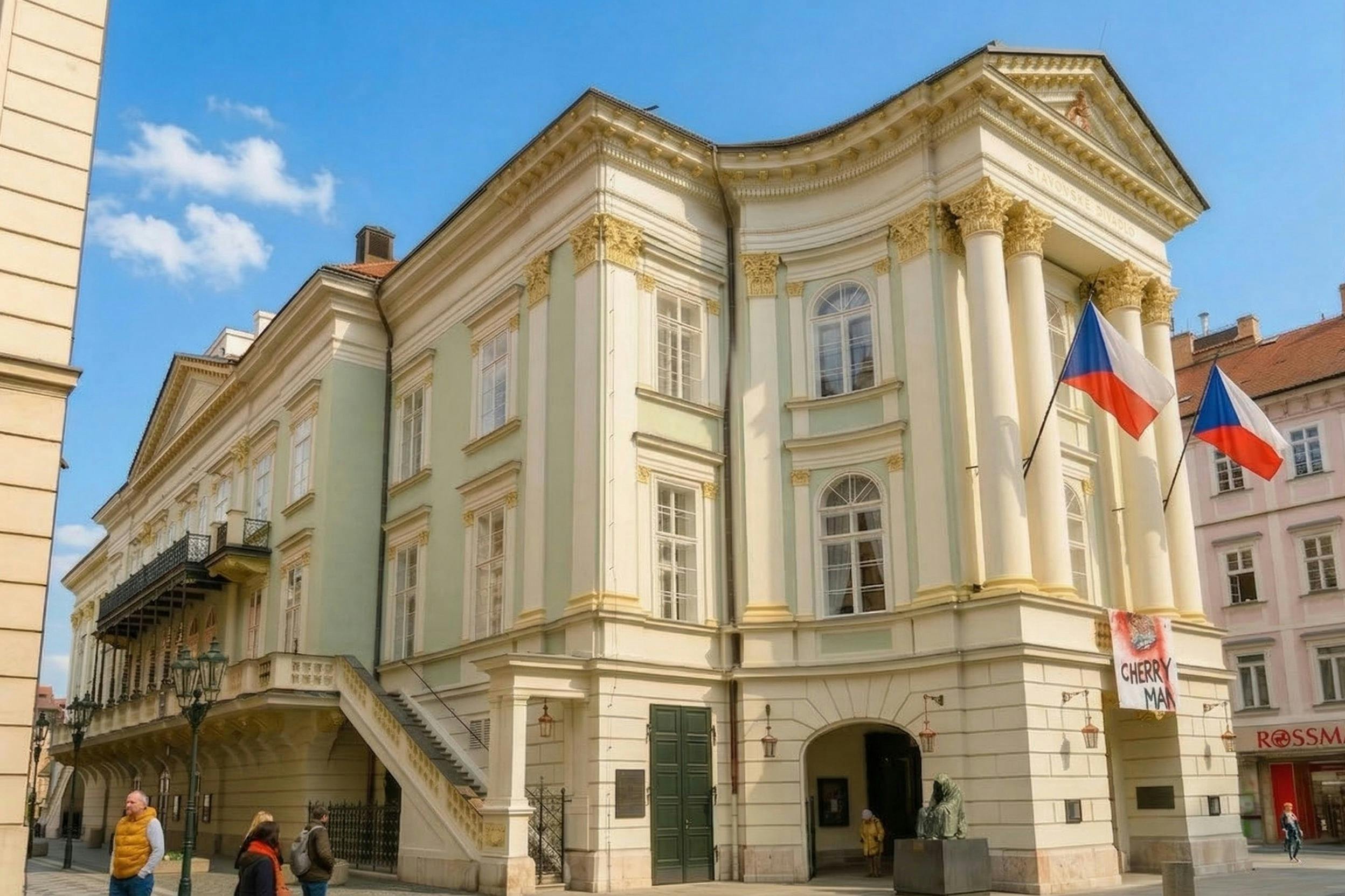 A light-colored historic building with columns, large windows, and a Czech flag. People are visible nearby in the scene.