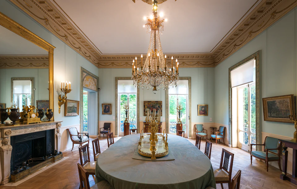 Elegant dining room with a large chandelier, an oval table with a decorative centerpiece, surrounded by chairs and paintings on the walls.