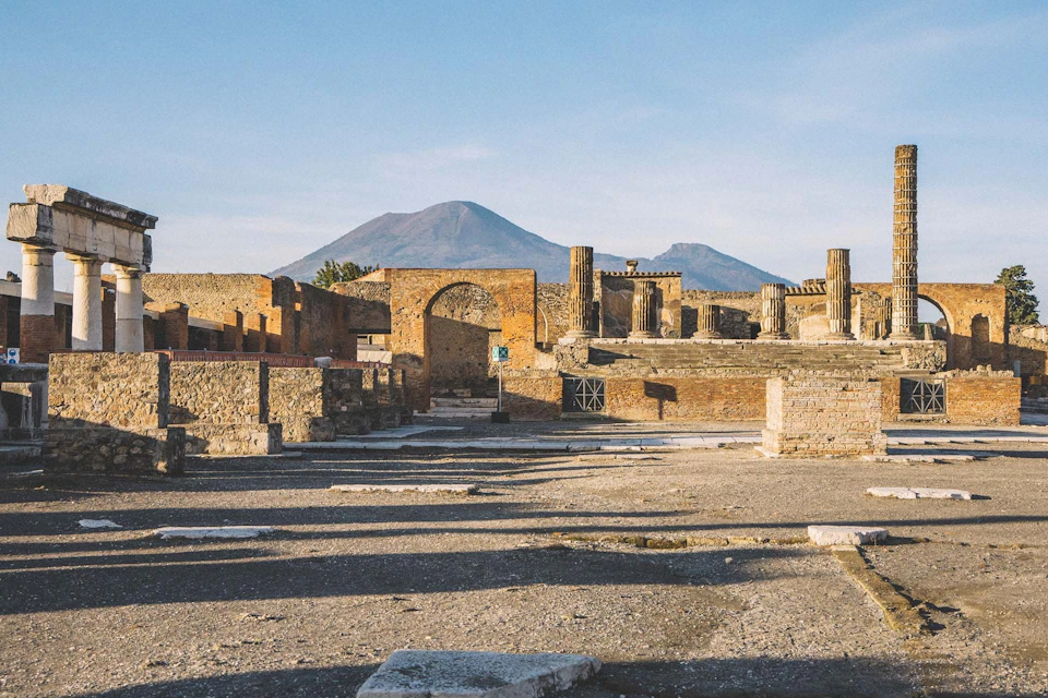Ancient ruins with stone columns and walls under a blue sky, with a mountain in the background.