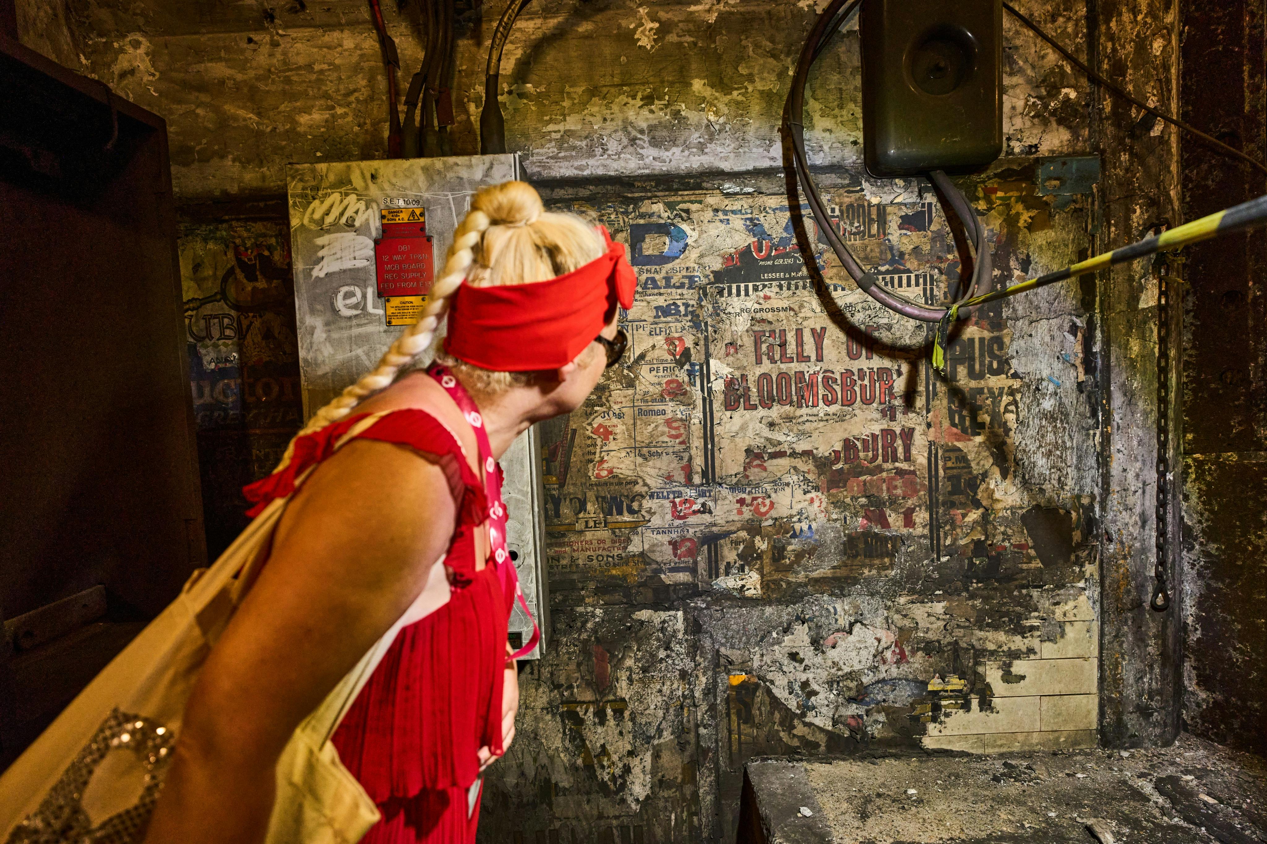 A person with a red dress and headband examines a weathered wall covered with vintage posters and exposed wiring in a dimly lit space.