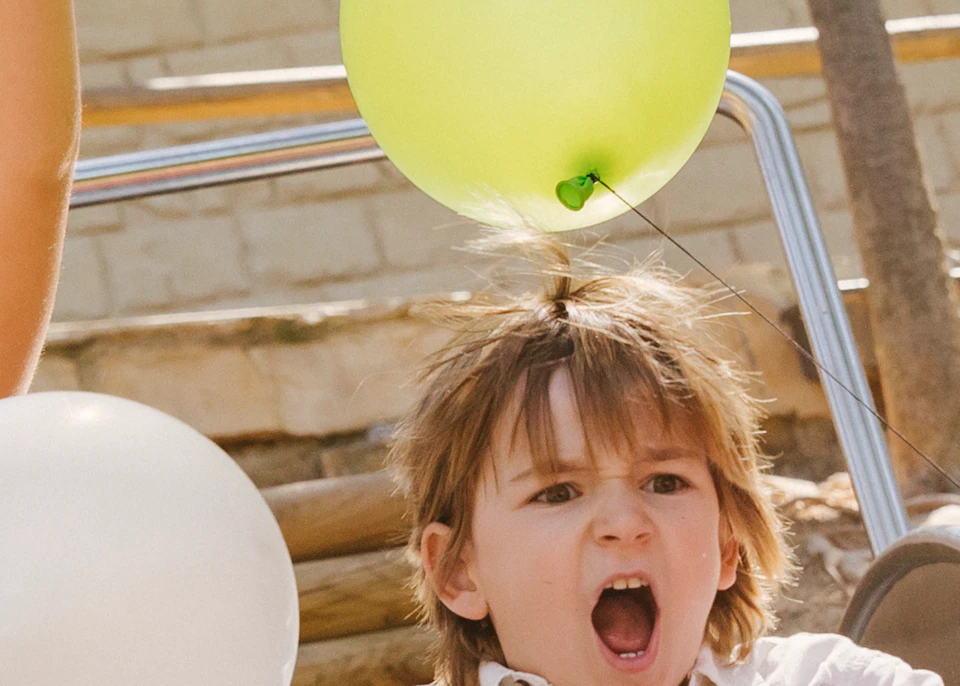 Three children holding balloons slide down a playground slide, laughing and looking excited.