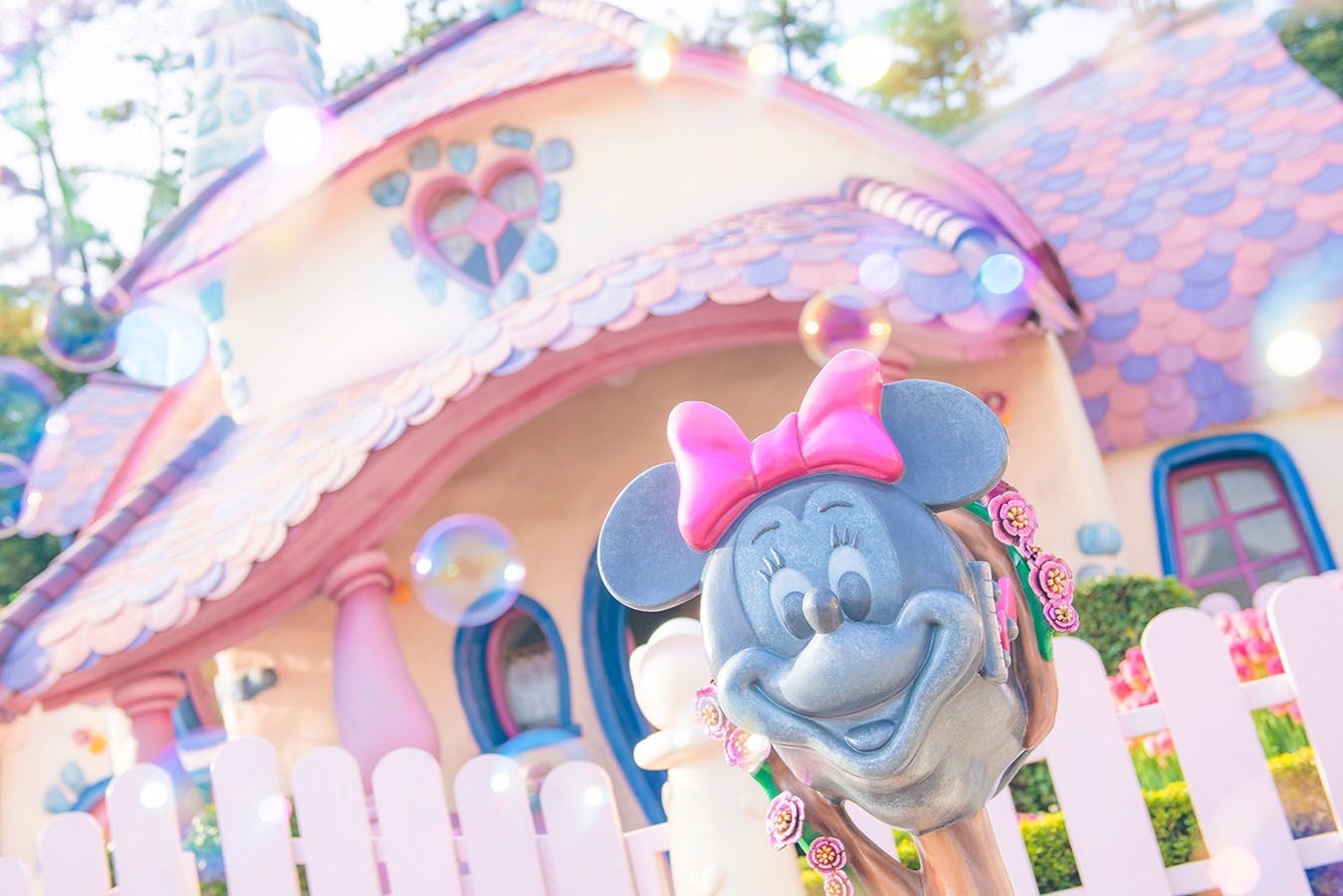 Ένα άγαλμα της Minnie Mouse με ροζ φιόγκο μπροστά από ένα ευφάνταστο σπίτι με διακόσμηση σε σχήμα καρδιάς και παστέλ χρώματα.