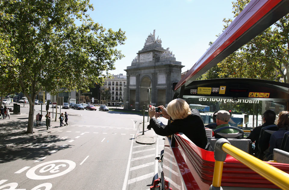 A person taking photos from the upper deck of a red tour bus, with a historic archway and city scene in the background.