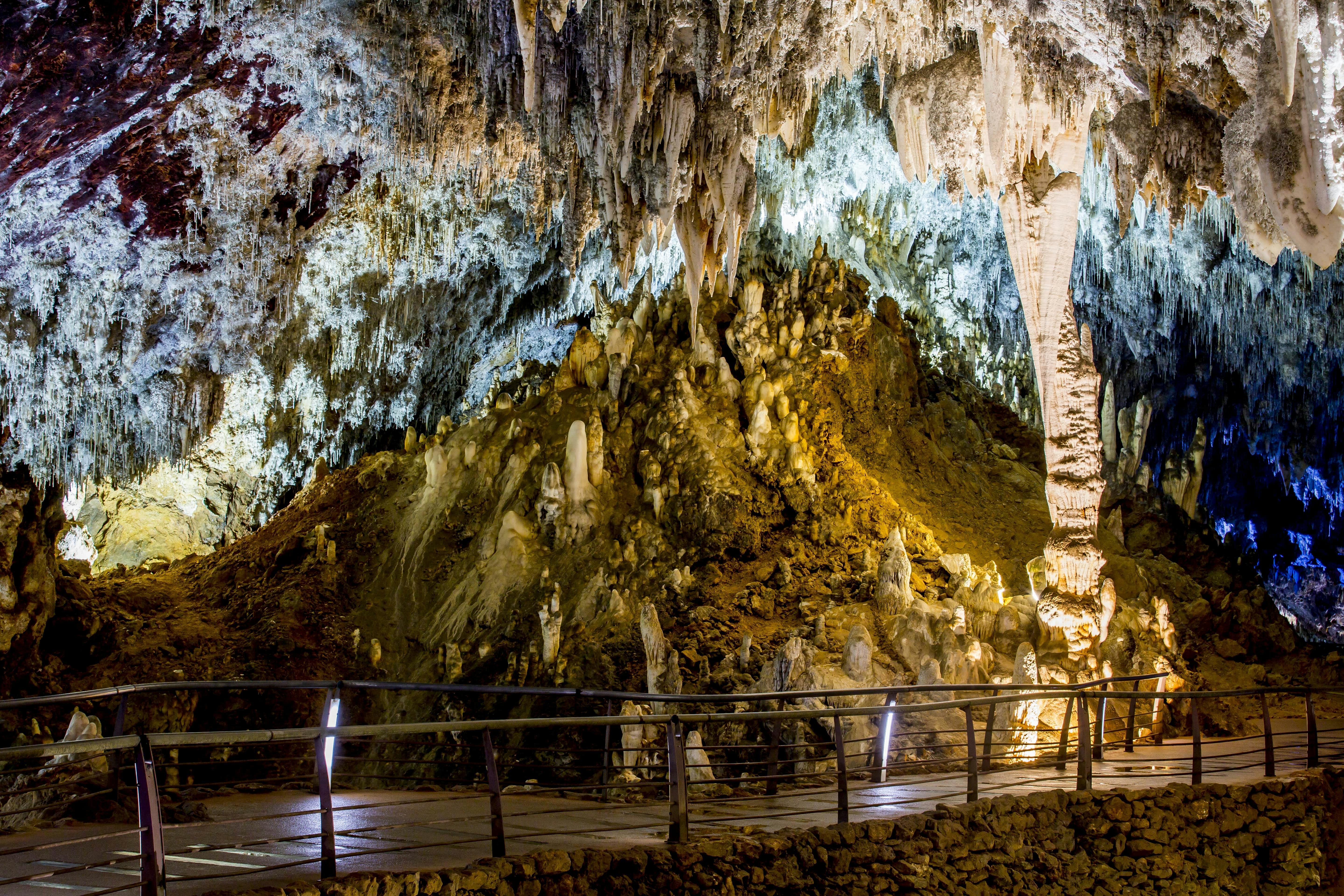 En hule med oplyste stalaktitter og stalagmitter, en gangbro i træ med rækværk i forgrunden.