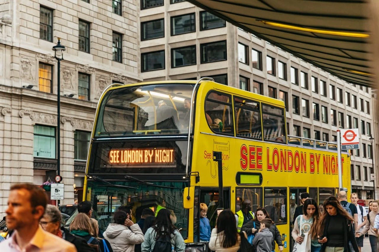 A yellow double-decker bus in a city labeled "See London By Night," surrounded by people and tall buildings.