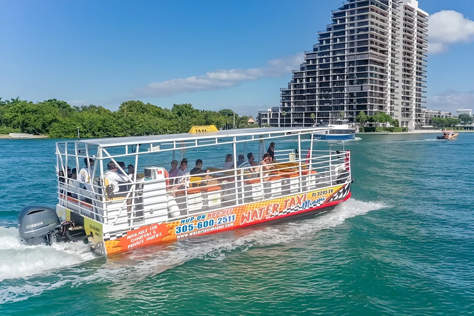 A water taxi with passengers cruises on a greenish-blue body of water, with a modern high-rise building and trees in the background.