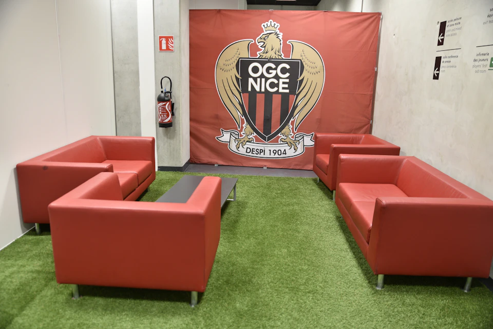 Four red armchairs and a small black table on green carpet with an OGC Nice banner in the background.