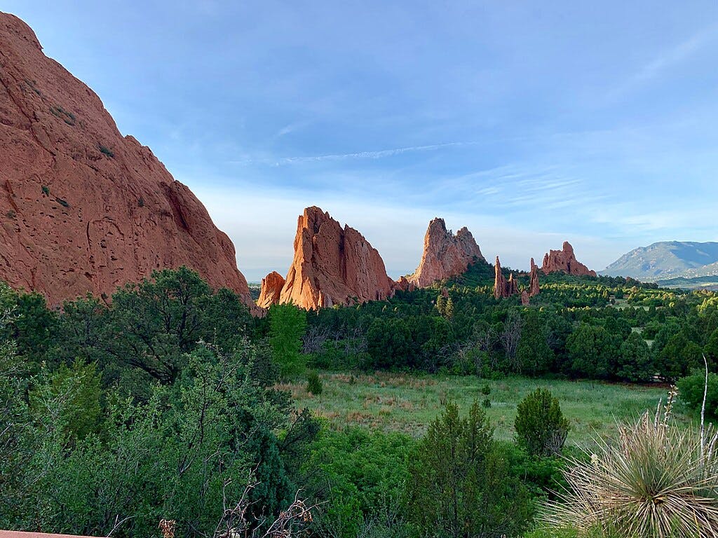 Dramáticas formações rochosas vermelhas erguem-se da vegetação verdejante sob um céu azul claro, com montanhas visíveis ao fundo.