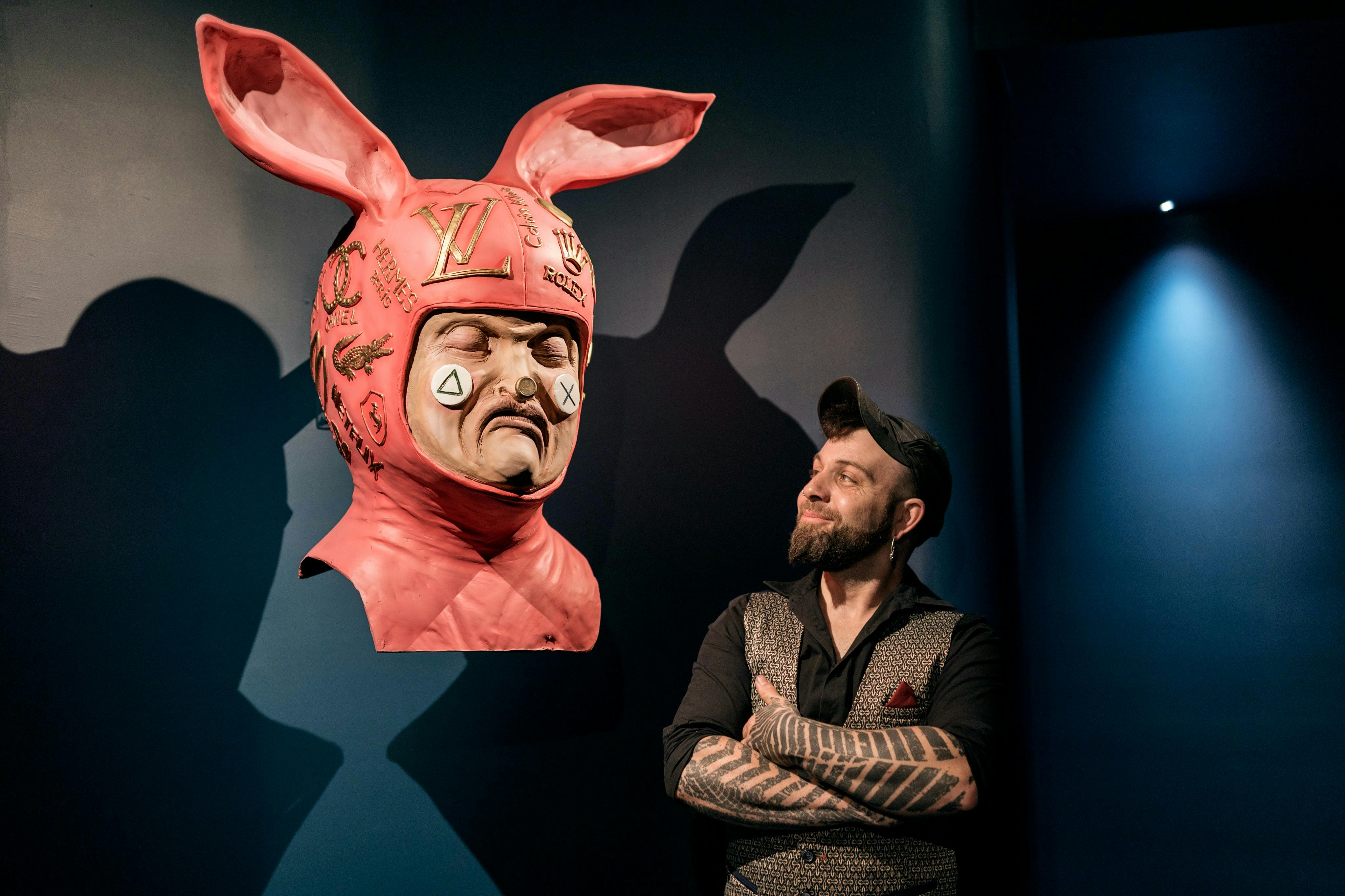 Un homme au bras tatoué regarde une sculpture de casque rose à oreilles de lapin avec divers logos sur un mur bleu dans une galerie d'art.