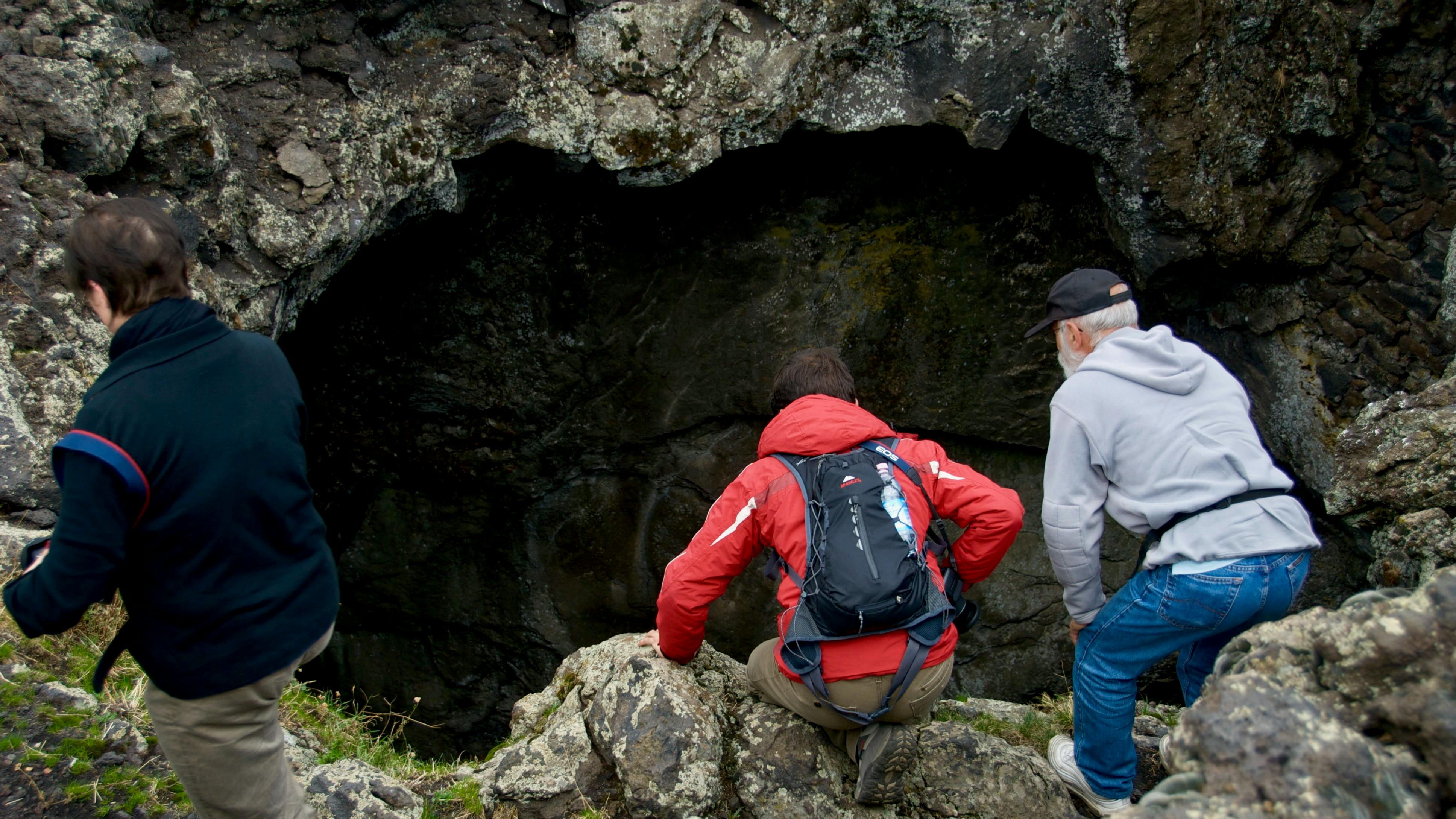 Drie mensen turen in een grote grotopening in een rotsachtig landschap. Eén persoon draagt een rode jas en een rugzak.