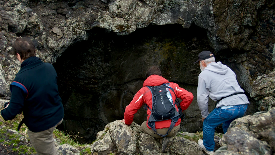 Three people are peering into a large cave opening in a rocky landscape. One person wears a red jacket and a backpack.