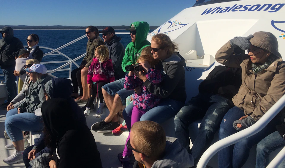 People on a boat are dressed warmly; one is using binoculars. The boat has "Whalesong" written on it.