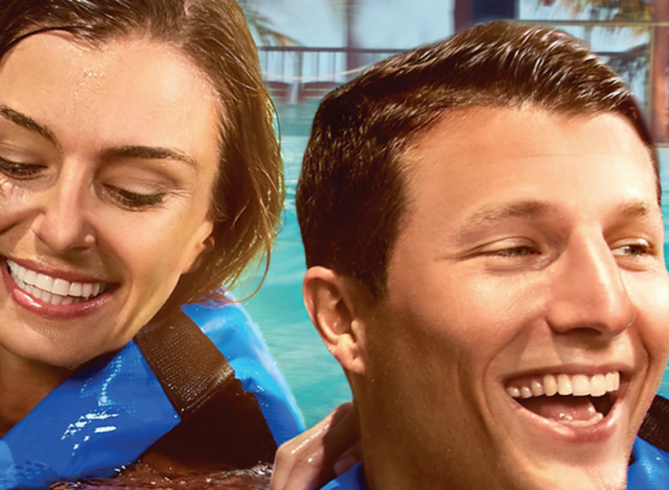 Four people in life vests smile while interacting with a dolphin in a pool, with palm trees and a wooden structure in the background.