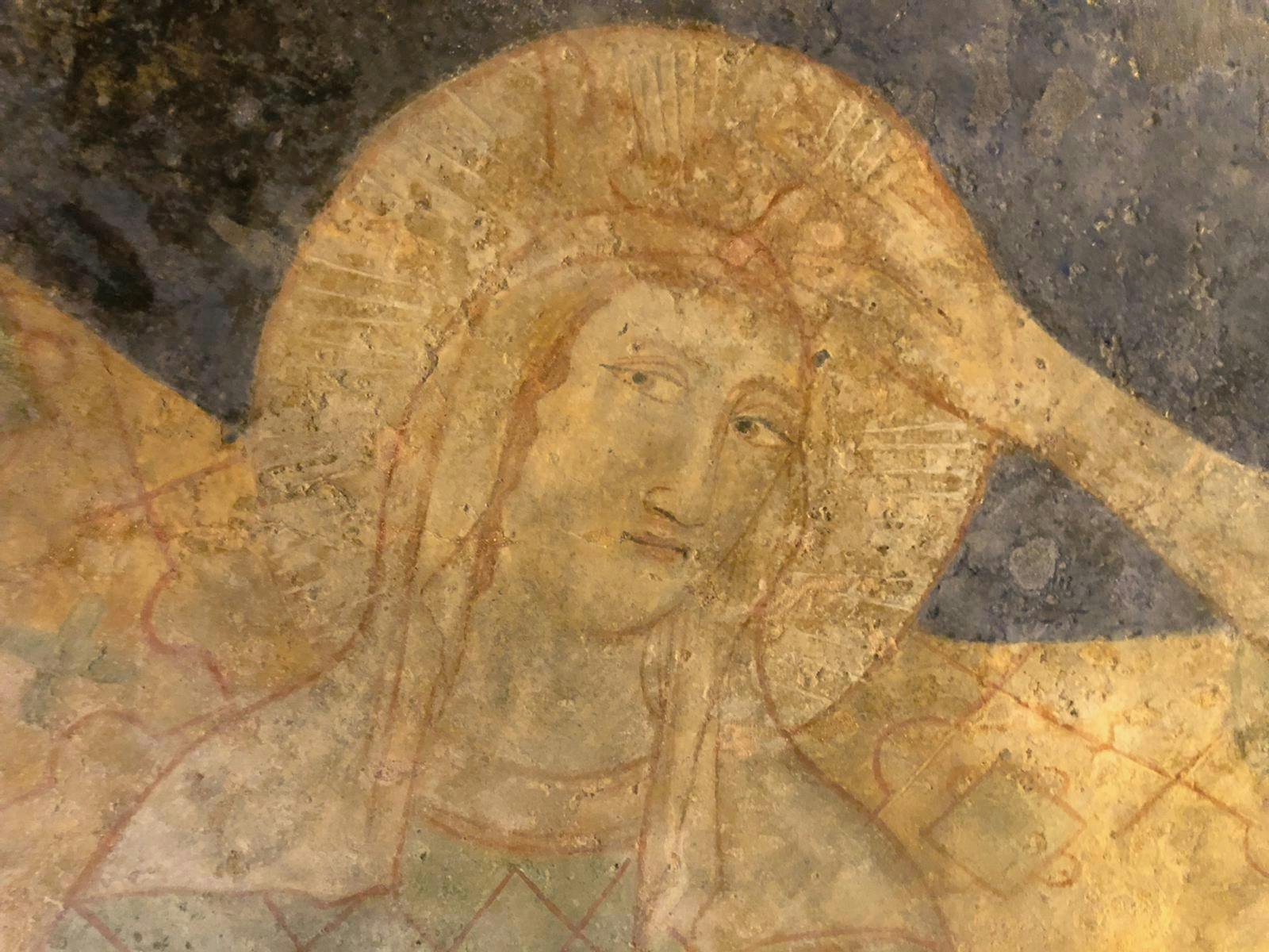 Starobylá freska zobrazující klidnou postavu s osvětlenou svatozáří, lehce opotřebovanou a posazenou na texturovaném, zvětralém pozadí.