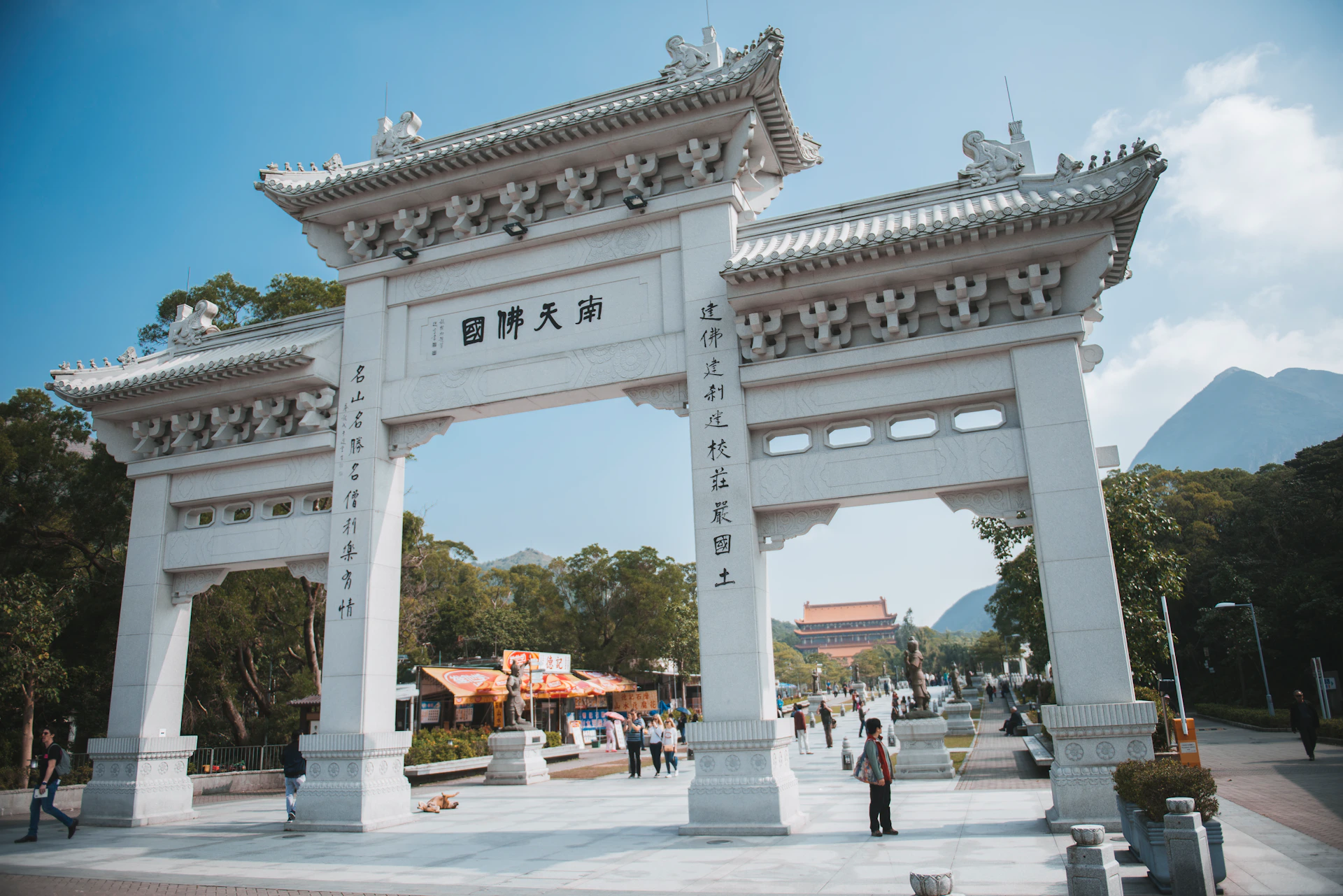White traditional Chinese archway with ornate designs. People walking and trees in the background under a clear blue sky.