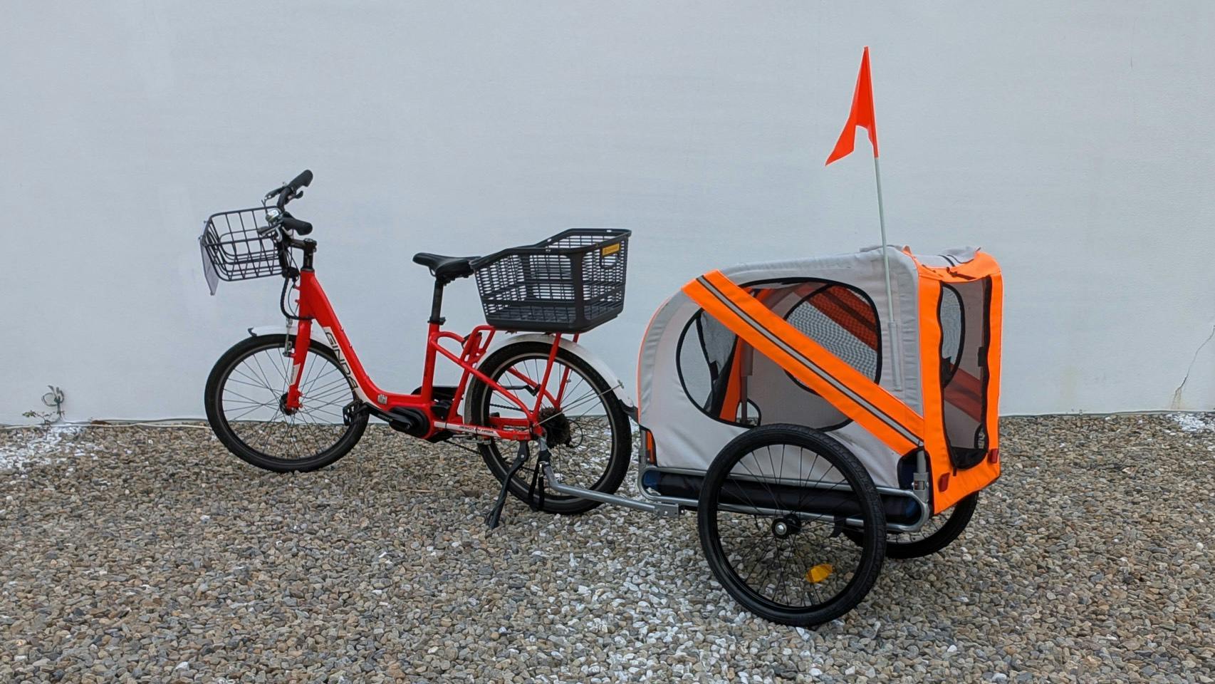 A red bicycle with a basket is attached to a gray and orange trailer with a flag, parked on a gravel surface.