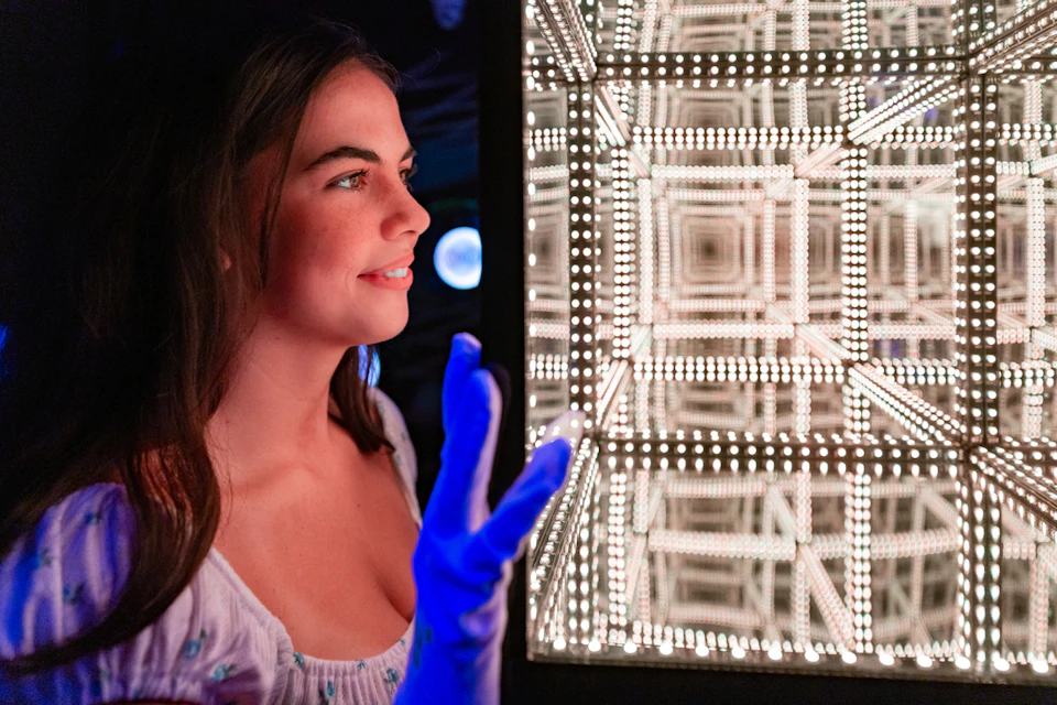 A person wearing a light-colored top and blue glowing gloves looks at a brightly lit mirrored structure in a dark setting.