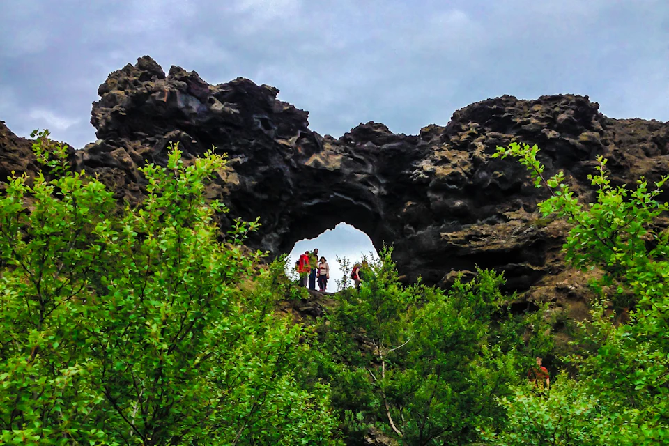 Three people stand on a rocky archway surrounded by lush greenery, with a cloudy sky in the background.