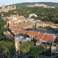 Flygfoto över en historisk stad med tegeltak, stenbyggnader och omgivande grönska, mot en kuliss av avlägsna kullar och stora strukturer.