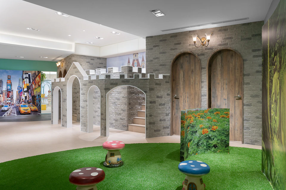 Indoor play area with castle-themed decor, mushroom stools on green carpet, and wooden doors in a brick wall.