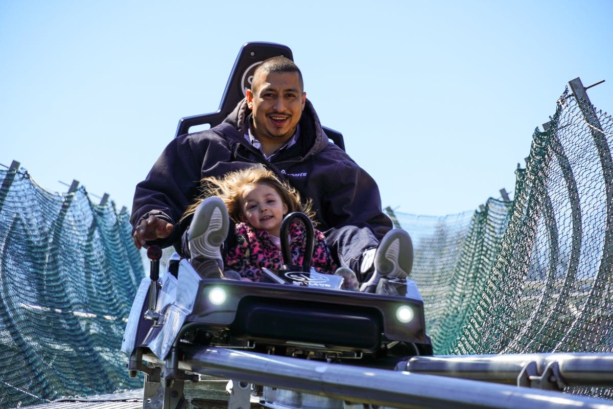 Ένας ενήλικας και ένα παιδί χαμογελούν ενώ οδηγούν ένα τρενάκι του λούνα παρκ. Το παιδί φοράει ροζ και περιβάλλεται από ένα δικτυωτό φράγμα.